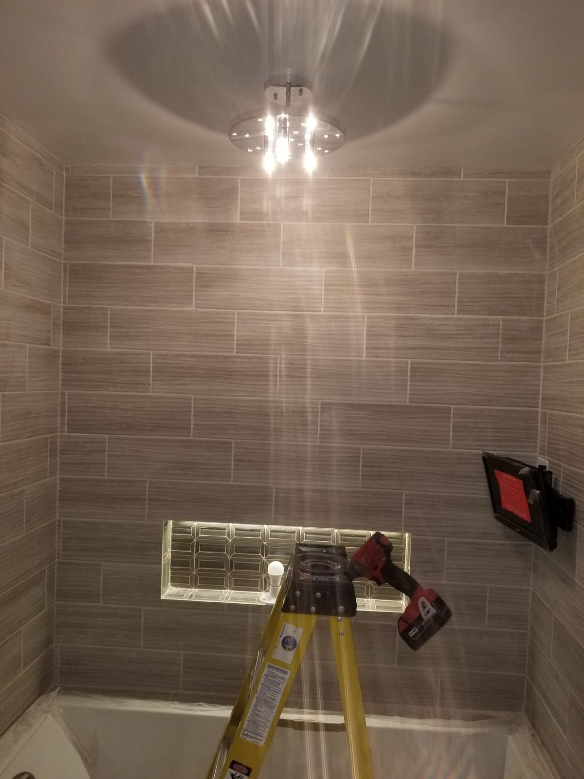 A new LED ceiling fixture lit up in a tiled bathroom, with the ladder still in place after installation.