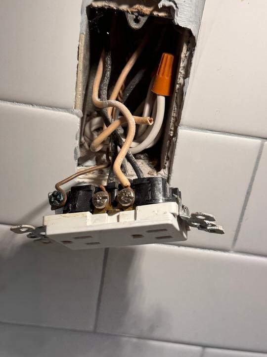 An old receptacle in a tiled wall with brittle insulation and crowded wiring, pulled out for replacement.